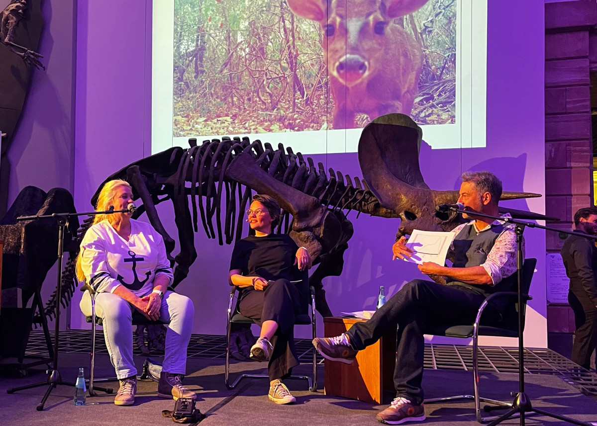 Drei Personen auf eine Bühne im Gespräch. Im Hintergrund das Skellett eines Dinosauriers.