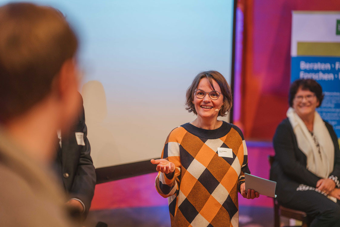 Susan auf einer Bühne, im Gespräch mit Menschen. Susan lächelt und übergibt das Gespräch mit einer Geste an einen Gesprächspartner am Rand des Bildes.