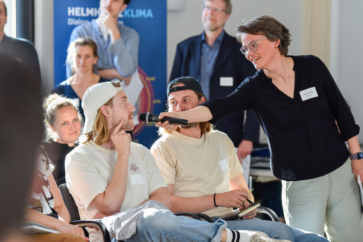 Susan auf der rechten Seite des Bildes, mit Mikrofon in der Hand. Auf Stühlen sitzen Personen, die Interessiert zuhören. Susan hält einem Mann das Mikrofon zu, der darein spricht.