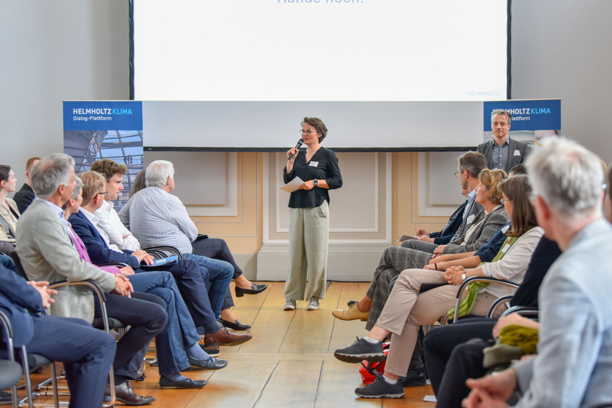 Susan steht in der Mitte des Bildes vor einem Präsentationsscreen, in der einen Hand hält sie ein Mikrofon in das sie spricht und in der anderen Hand Blätter mit Notizen. Links und Rechts sitzen Menschen und hören zu. Auf zwei Bannern steht in weiß auf bau 'HELMHOLTZ KLIMA'.